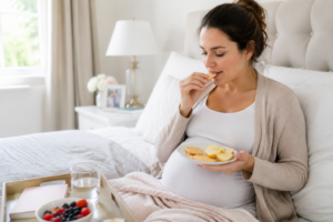 Pregnant woman eating small healthy snack (crackers, fruit) beside bed in morning